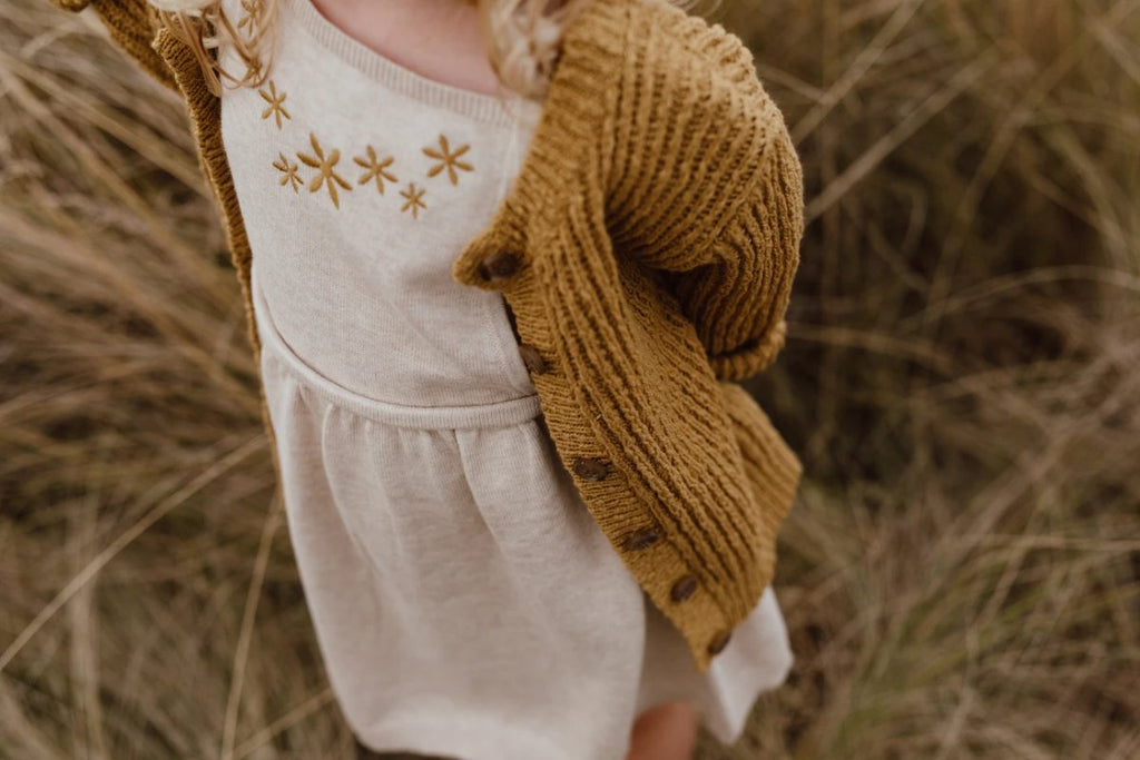 US stockist of Grown Clothing's Flower Field dress made from organic cotton.  Features gathered skirt, ties at shoulder and had machine embroidered gold flowers on the bib.  Oatmeal in color.