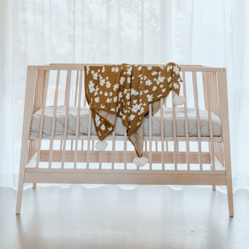 US stockist of Grown Clothing's gender neutral, organic cotton Winter Wonder Pom Pom Baby Blanket in bronze.  Made from organic cotton, with contrasting white jacquard flowers and white pom poms on each corner.