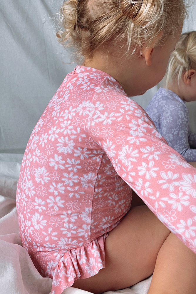 US stockist of Salty Swimwear's Zoe long sleeve rashsuit in pink daisy days.  Made from pink UPF 50+ recycled Repreve fabric with pretty white daisy print. Fully lined, with invisible back zipper and signature Salty frill on legs.