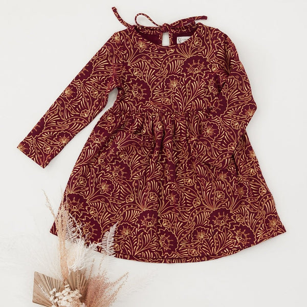 US stockist of Karibou Kids' Royal Visit Long Sleeve Pocket Dress in Cherry Red.  Made from cotton in a rich cherry red color with contrasting gold swirl print and ties at neck.